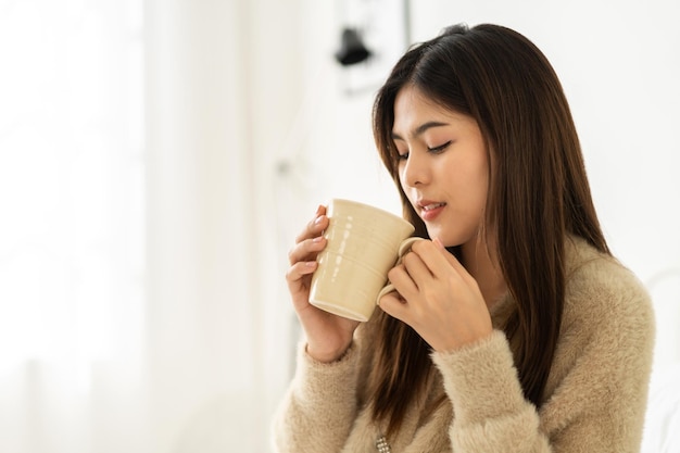 Retrato de una mujer asiática sonriente, feliz y alegre, que se relaja bebiendo y mirando una taza de café caliente o té