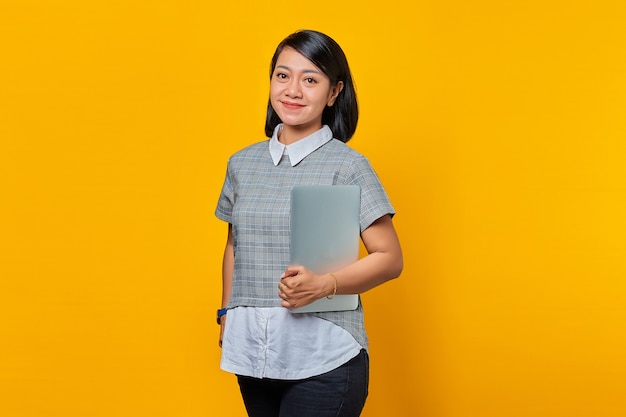 Retrato de mujer asiática sonriente traer portátil sobre fondo amarillo