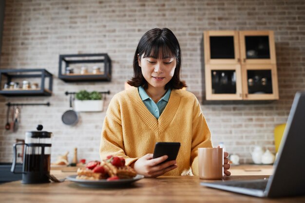 Retrato de una mujer asiática sonriente usando un teléfono inteligente mientras disfruta del desayuno en el acogedor espacio de copia de la cocina casera