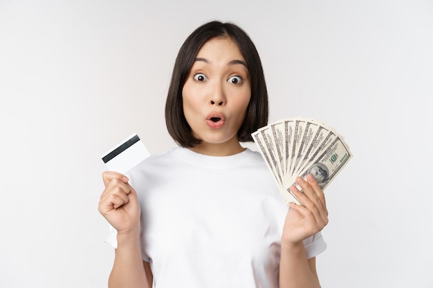 Retrato de mujer asiática sosteniendo dólares de dinero y tarjeta de crédito que parece impresionada y asombrada de pie en camiseta sobre fondo blanco.