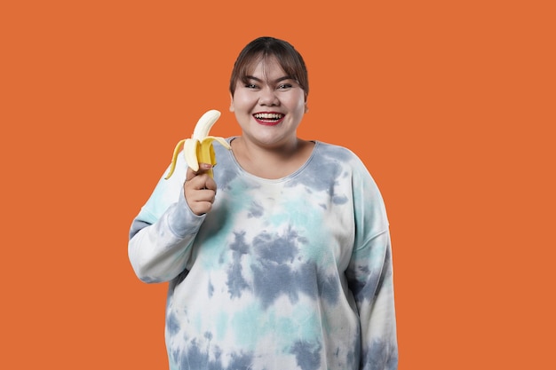 Retrato de mujer asiática sosteniendo plátano, aislado sobre fondo naranja, concepto de nutrición saludable