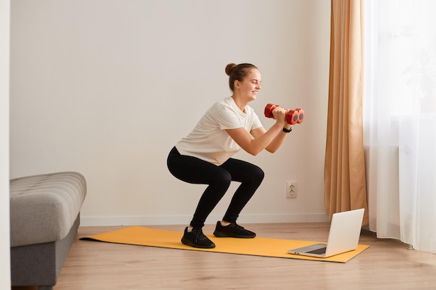 Retrato de una mujer atlética con moño de pelo haciendo ejercicio deportivo en cuclillas, en la parte inferior del cuerpo, manteniendo el equilibrio, calentando y entrenando los músculos, sosteniendo en las manos un tonto rojo, usando una camiseta blanca y leggins.