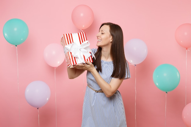 Retrato de mujer atractiva alegre en vestido azul mirando caja roja con regalo presente sobre fondo rosa pastel con coloridos globos de aire. Fiesta de cumpleaños, concepto de emociones sinceras de personas.