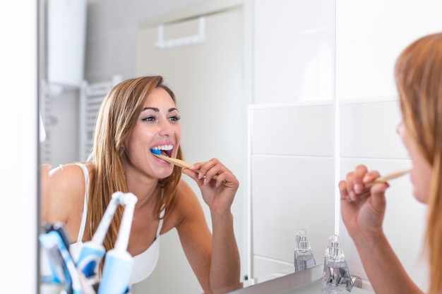 Retrato de una mujer atractiva cepillándose los dientes en el baño y mirándose en el espejo con dientes sanos en el reflejo Una joven encantadora y feliz mirándose al espejo y cepilándose los ojos en el baño