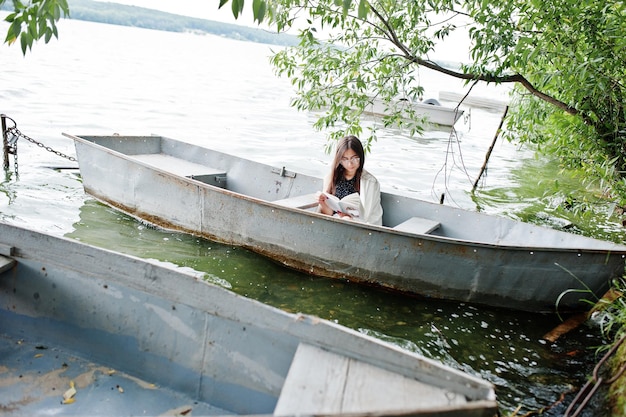 Retrato de una mujer atractiva con lunares negros, chal blanco y gafas leyendo un libro en un bote en un lago