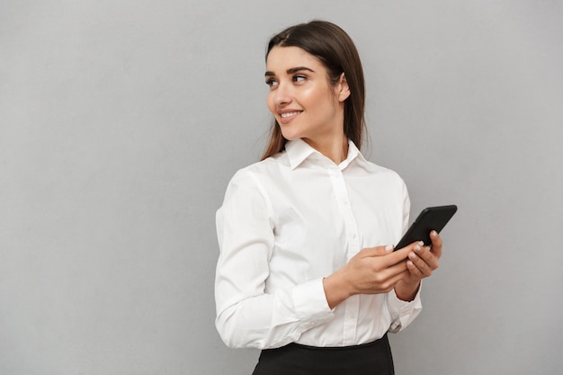 Retrato de mujer atractiva de negocios con cabello largo castaño en ropa formal sonriendo y mirando a un lado mientras sostiene el teléfono celular, aislado sobre la pared gris