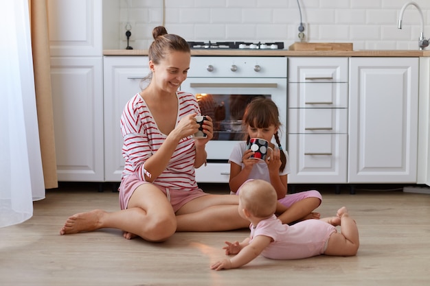Retrato de mujer atractiva con la prohibición del cabello vistiendo camisa casual a rayas sentada en el piso en la cocina con la taza en las manos, pasando tiempo con sus pequeñas hijas.