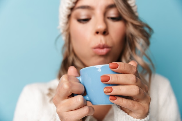 Retrato de mujer atractiva rubia con sombrero blanco soplando y sosteniendo la taza