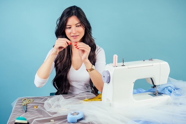 Retrato mujer atractiva sastre modista (costurera) enhebrar la aguja en la máquina de coser sobre un fondo azul en el estudio. El concepto de crear una nueva colección de ropa.