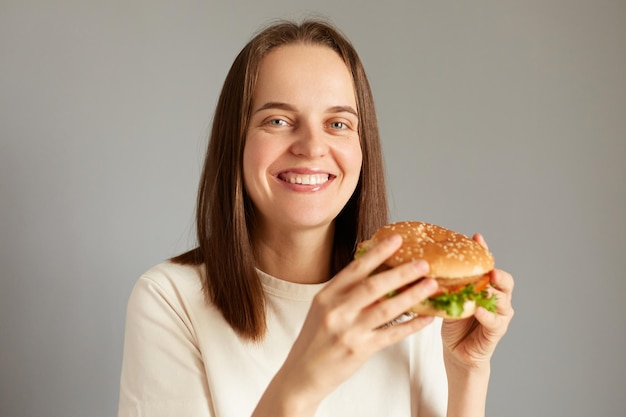 Retrato de una mujer atractiva sonriente que sostiene una deliciosa hamburguesa fresca comiendo un bocadillo de comida chatarra mira a la cámara con una expresión de alegría con una comida trampa disfrutando de un sabroso sándwich