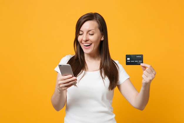 Retrato de una mujer bastante joven con ropa informal blanca usando un teléfono móvil, sosteniendo una tarjeta de crédito aislada en un fondo de pared naranja amarillo en el estudio. Concepto de estilo de vida de las personas. Simulacros de espacio de copia.