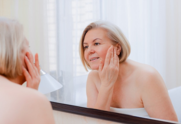 Retrato mujer bastante mayor con las manos en el espejo de la cara en casa después del baño Cuidado de la piel después de 50-60 años