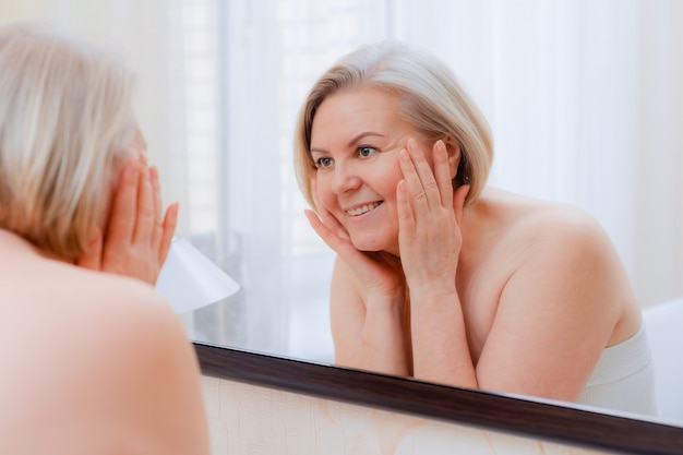 Retrato mujer bastante mayor con las manos en el espejo de la cara en casa después del baño Cuidado de la piel después de 50-60 años