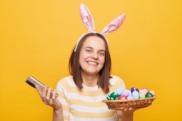 Retrato de una mujer bastante satisfecha con orejas de conejo sosteniendo huevos de Pascua y un teléfono inteligente mirando la cámara con una sonrisa con dientes usando un teléfono aislado en un fondo amarillo