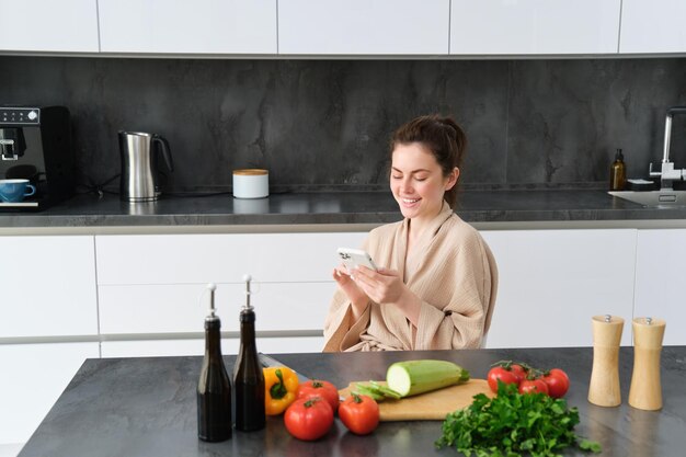 Retrato de una mujer en bata de baño sentada en la cocina con un teléfono inteligente cocinando la cena viendo la receta