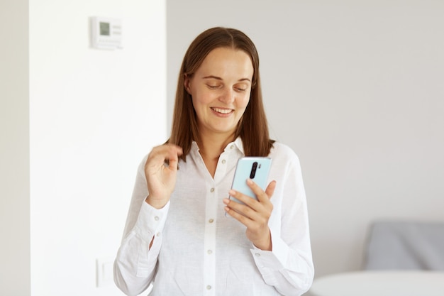 Retrato de mujer blogger optimista con camisa blanca de estilo casual, posando en una habitación luminosa en casa, usando un teléfono inteligente para tomar autofotos o transmitir en vivo.