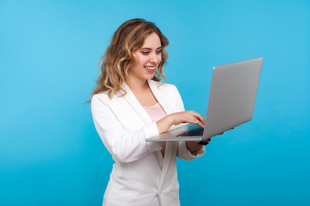 Retrato de mujer bonita alegre con cabello ondulado en chaqueta blanca de pie con mensaje de escritura de computadora portátil y sonriendo genuinamente charlando en la red social tiro de estudio interior aislado sobre fondo azul