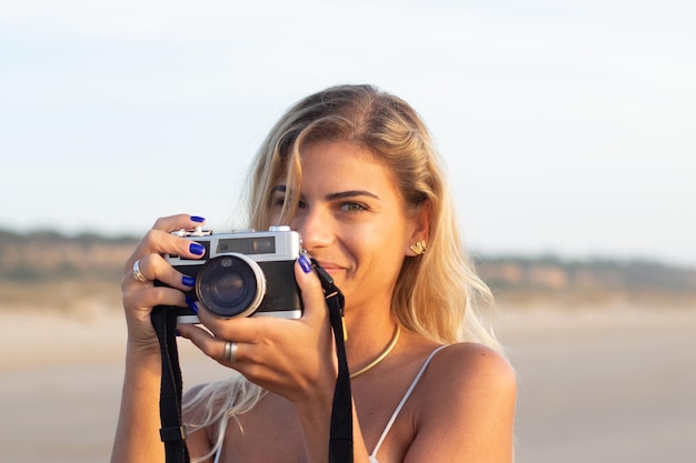 Retrato de mujer bonita con cámara antigua. Modelo femenino con cabello rubio y ropa informal sosteniendo una cámara en la playa. Hobby, concepto de viaje