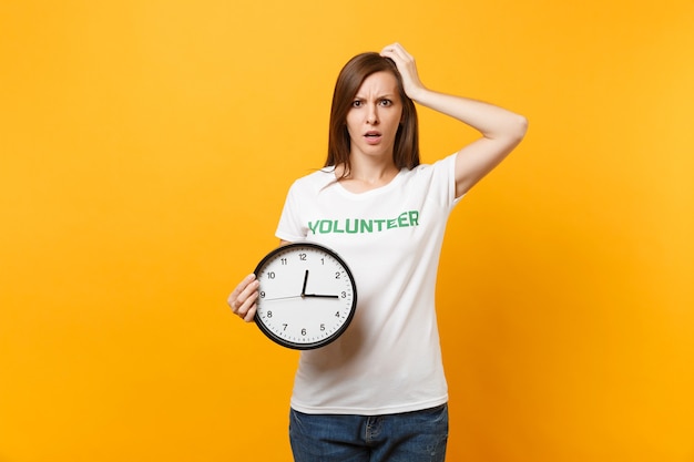 Retrato de mujer bonita en camiseta blanca con inscripción escrita voluntario de título verde mantenga reloj redondo aislado sobre fondo amarillo. Ayuda de asistencia gratuita voluntaria, concepto de tiempo de trabajo de gracia de caridad