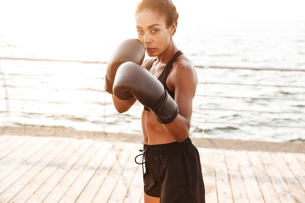 Retrato de mujer bonita enfocada vistiendo ropa deportiva entrenamiento en guantes de boxeo junto al mar en la mañana
