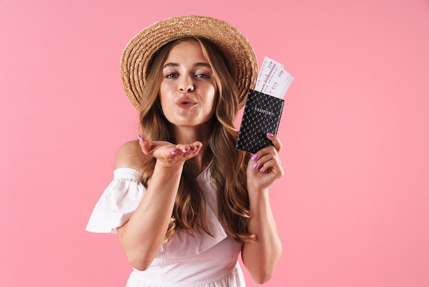 Retrato de una mujer bonita joven complacida posando aislada sobre pared rosa con pasaporte con boletos soplando besos.