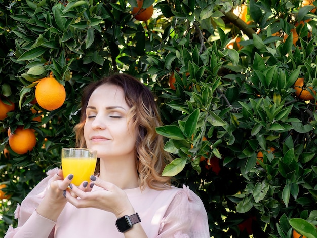 Retrato de mujer bonita joven sosteniendo un vaso de jugo de naranja cerca de naranjo