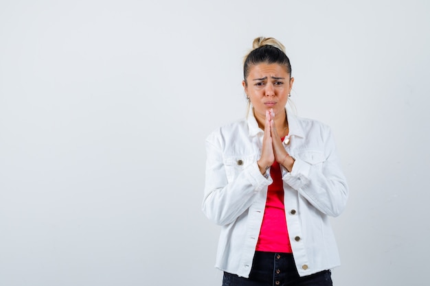 Retrato de mujer bonita manteniendo las manos en gesto de oración en chaqueta blanca y mirando triste vista frontal