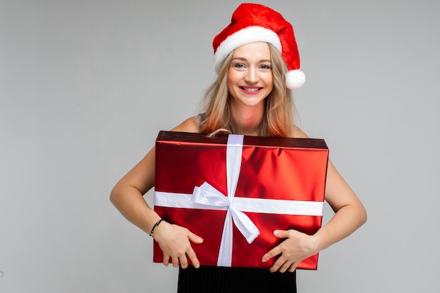 Retrato de mujer bonita rubia con sombrero rojo de Santa abrazando envuelto regalo de Navidad en papel de regalo rojo y lazo blanco. Chica rubia sonriendo a la cámara con regalo en las manos.