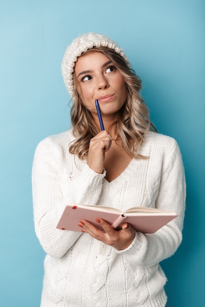 Retrato de mujer bonita soñando con gorro de lana mirando hacia arriba y haciendo notas en el diario