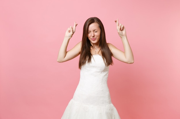 Retrato de mujer bonita en vestido blanco esperando un momento especial manteniendo los dedos cruzados, los ojos cerrados