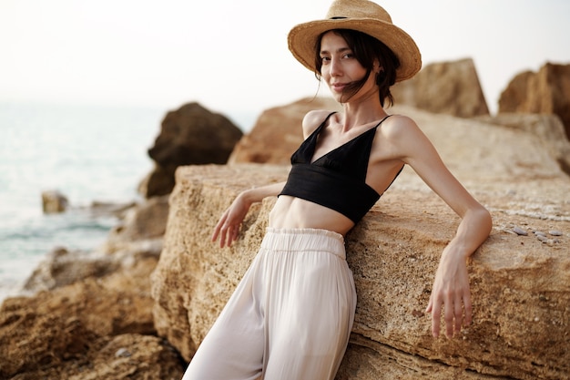 Retrato de mujer en bralette negro y pantalón blanco apoyado sobre una roca en la playa