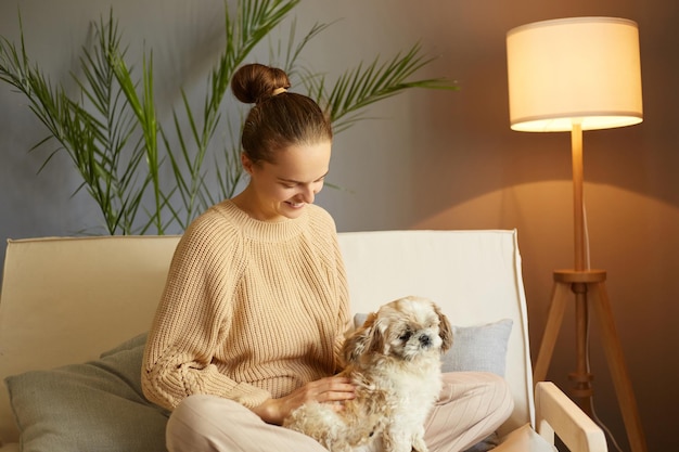 Retrato de una mujer de cabello castaño con suéter cálido acariciando a su amado perro pequinés sentado en el sofá pasando tiempo el fin de semana con su animal favorito