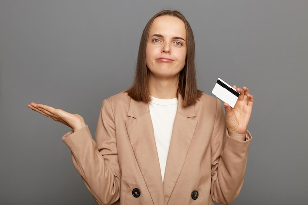 Retrato de una mujer de cabello castaño triste e insatisfecha que usa una chaqueta que sostiene una tarjeta de crédito de plástico y muestra un gesto de no saber encogiéndose de hombros posando aislado sobre un fondo gris