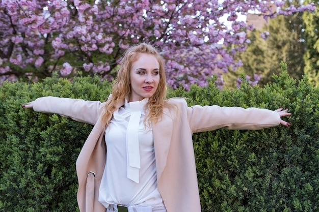 Retrato de una mujer de cabello escarlata con cabello rizado con los brazos levantados en un parque de primavera en flor Niña en un árbol de sakura en flor