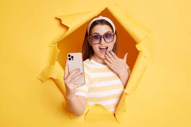 Retrato de una mujer de cabello oscuro sorprendida con camisa a rayas y banda para el cabello posando en un agujero de papel amarillo sosteniendo un teléfono inteligente y cubriendo la boca con la palma ve algo asombrado