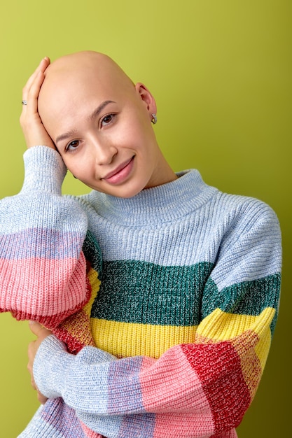 Retrato de una mujer calva caucásica sonriente con una camisa de colores brillantes tocando la cabeza posando aislada sobre el fondo verde del estudio. La dama atractiva tiene la enfermedad de alopecia. Salud, belleza, gente.