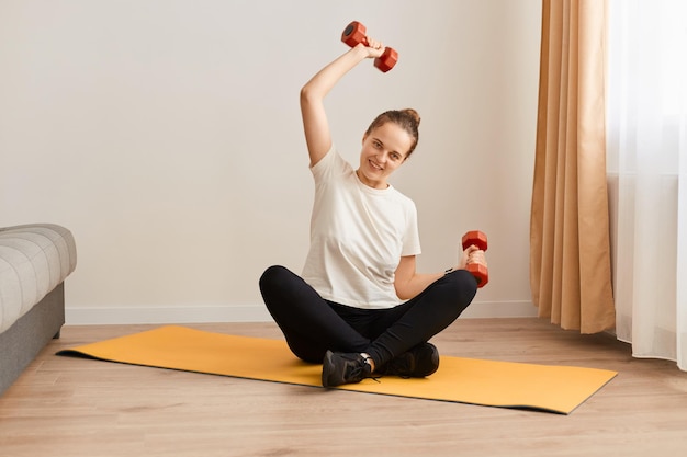 Retrato de mujer con camiseta blanca y leggins negros haciendo ejercicio con pesas en casa entrenando bíceps y tríceps ejercicio físico vida sana y dieta