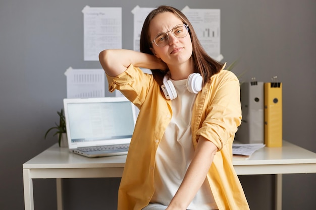 Retrato de una mujer cansada y exhausta con cabello castaño y camisa amarilla posando en la oficina con exceso de trabajo sufriendo dolor en el cuello frunciendo el ceño