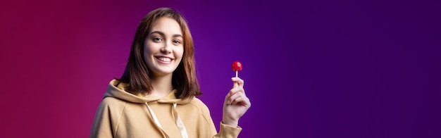 Retrato de una mujer con capucha lamiendo una piruleta redonda roja con un hermoso maquillaje en un fondo morado de moda en el estudio