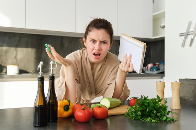 Retrato de mujer con cara enojada parada cerca de verduras y luciendo frustrada sosteniendo un cuaderno