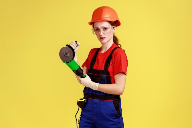 Retrato de una mujer carpintera seria que trabaja con una amoladora angular, con una expresión facial segura, usando overoles y casco protector. Disparo de estudio interior aislado sobre fondo amarillo.