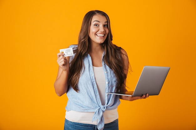 Retrato de una mujer casual joven feliz, sosteniendo la computadora portátil, mostrando tarjeta de crédito plástica