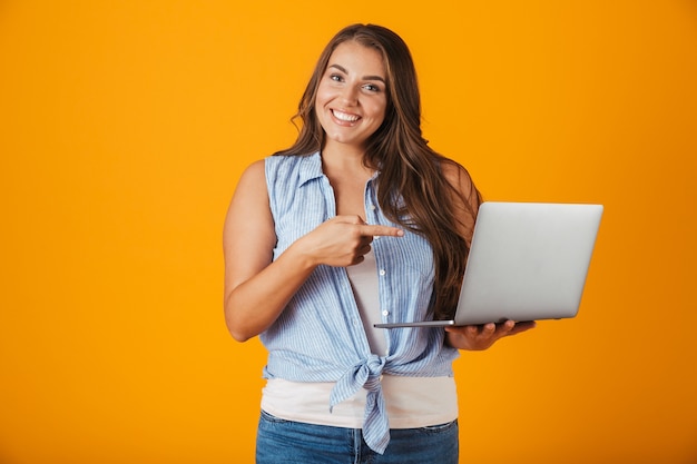 Retrato de una mujer casual joven feliz, sosteniendo la computadora portátil, señalar con el dedo