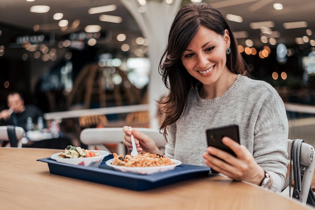 Retrato de una mujer casual positiva que come el almuerzo y que manda un SMS en el teléfono elegante.