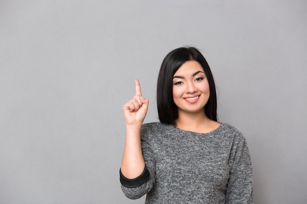 Retrato de una mujer casual sonriente apuntando con el dedo hacia arriba en la pared gris