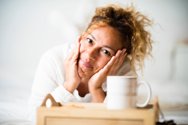 Retrato de mujer caucásica adulta joven hermosa alegre se acostó en el dormitorio en la cama con una taza de té o café - gente feliz en casa disfruta del concepto de despertar por la mañana