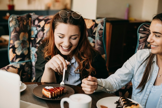 Retrato de una mujer caucásica alegre sentada en un café contando historias con su amiga mientras come tarta de queso y bebe café.