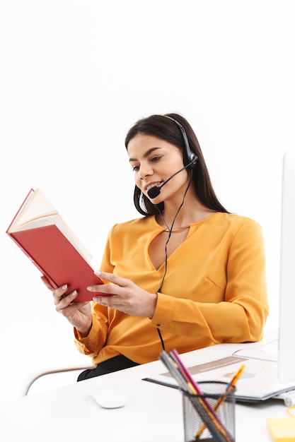 Retrato de mujer caucásica asistente de línea directa vistiendo auriculares con micrófono libro de lectura mientras trabaja en la oficina