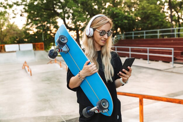 Retrato de mujer caucásica complacida usando audífonos y gafas de sol escribiendo en el teléfono celular mientras sostiene la placa en el skatepark