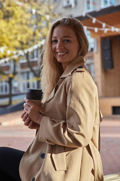 Retrato de mujer caucásica en gabardina beige al aire libre con una taza de café